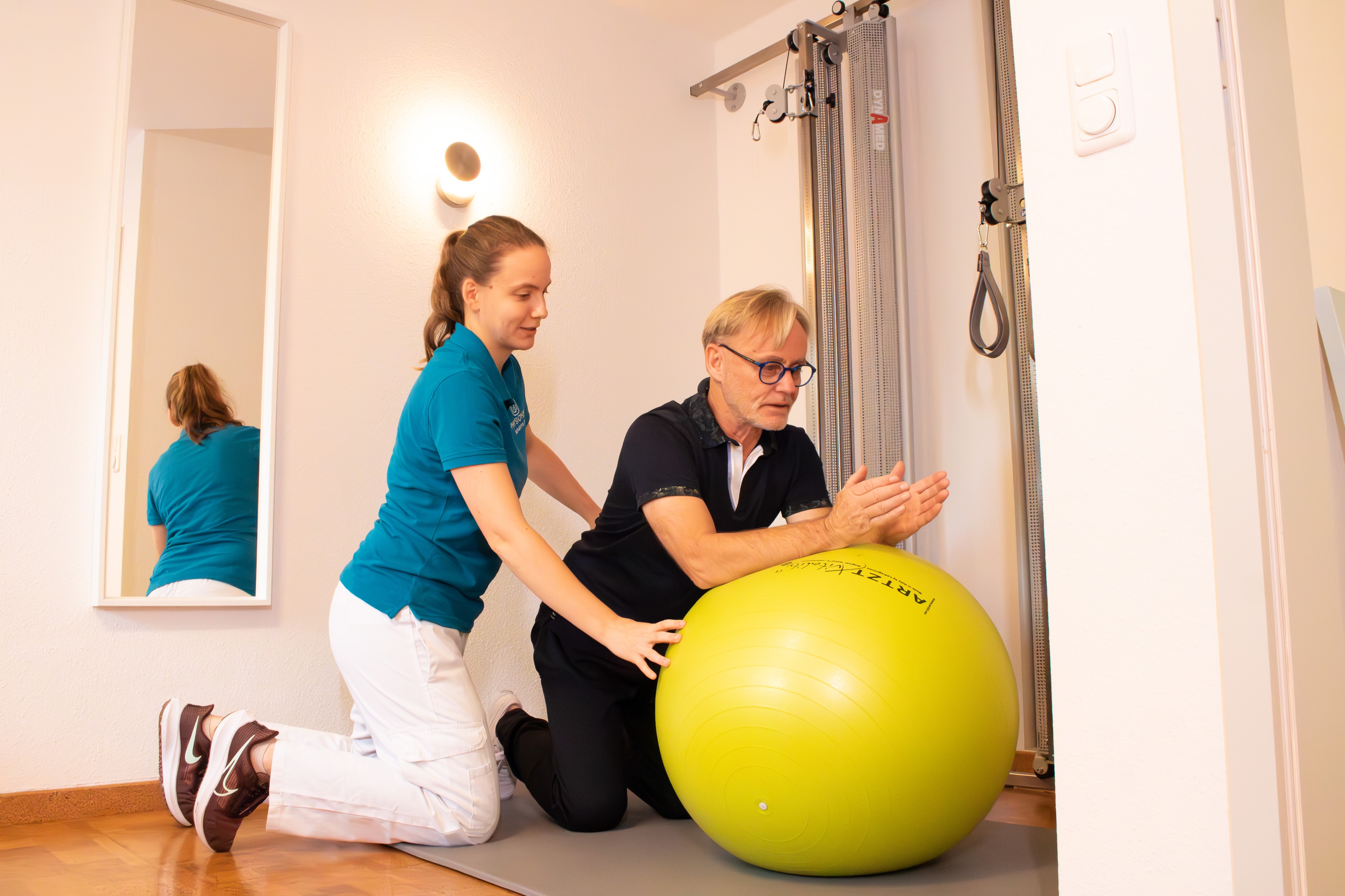 Physiotherapeutin Julia Neelsen steht hinter einer Patientin während diese leicht gebeugt eine Übung der Krankengymnastik macht.